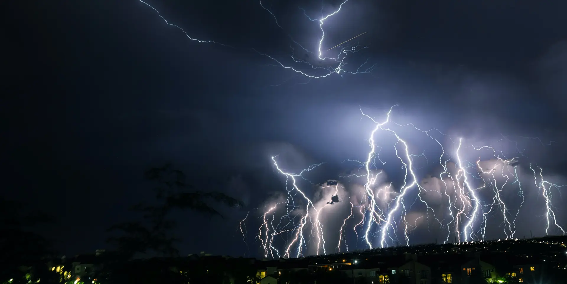 A smattering of white lightning bolts in a dark blue, night sky. Several houses with lit windows are in the the foreground.