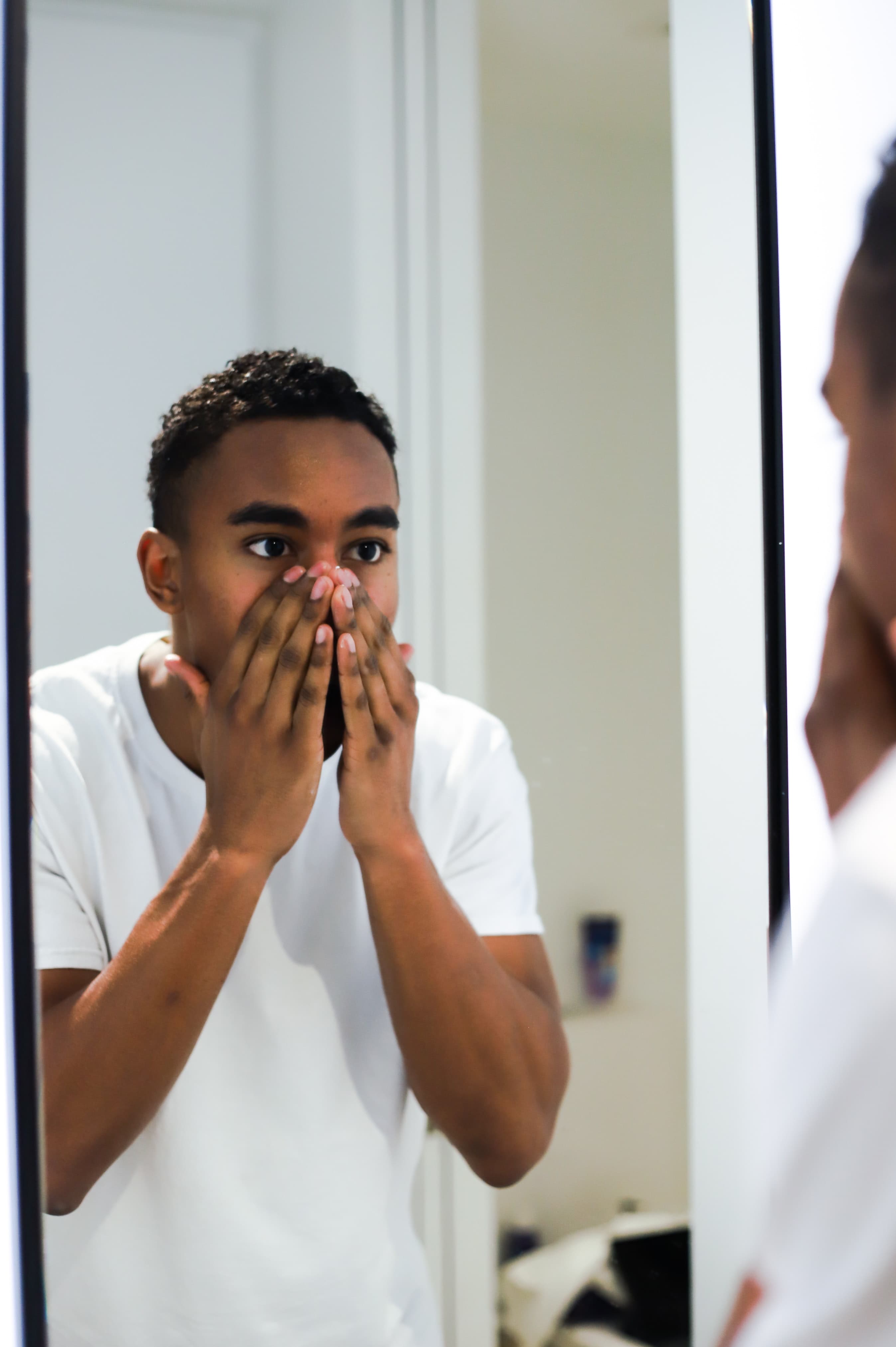 A young Black man looks at his reflection in the mirror. His hands are covering his nose, mouth, and chin.