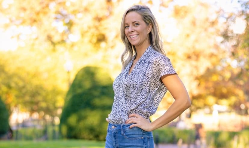 A smiling blonde woman in jeans and a short sleeve blouse smiles at the camera in front of a blurred background of trees and grass.